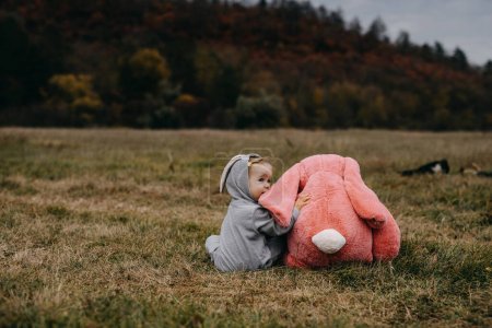 Foto de Little child wearing a bunny costume, sitting with a big pink plush bunny toy, hugging it, outdoors, in an open field. - Imagen libre de derechos