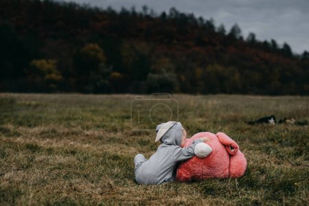 Téléchargez les photos : Petit enfant portant un costume de lapin, assis avec un gros lapin en peluche rose jouet, étreignant, à l'extérieur, dans un champ ouvert. - en image libre de droit