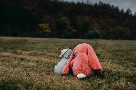 Foto de Little child wearing a bunny costume, playing with a big pink plush bunny toy, hugging it, outdoors, in an open field. - Imagen libre de derechos