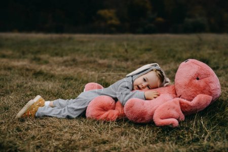 Foto de Little child wearing a bunny costume, playing with a big pink plush bunny toy, hugging it, outdoors, in an open field. - Imagen libre de derechos