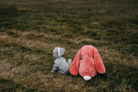 Foto de Little child wearing a bunny costume, sitting with a big pink plush bunny toy, outdoors, in an open field. - Imagen libre de derechos