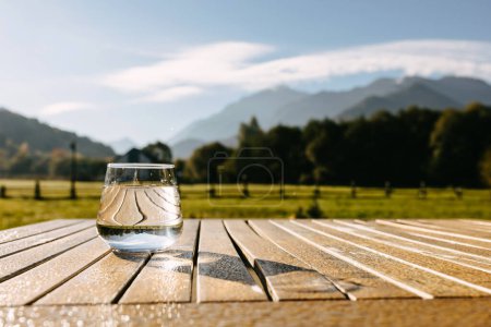 Photo for Glass of still water placed on a wooden table outdoors, in morning light, on mountains background. - Royalty Free Image