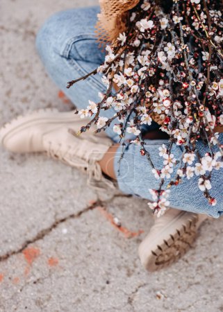 Photo for Closeup of a woman sitting on the floor in blue jeans, holding a big bunch of branches with pink cherry flowers in bloom. - Royalty Free Image