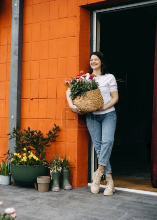 Photo for Florist working at a flower shop. Woman a big basket with pink flowers, smiling. - Royalty Free Image