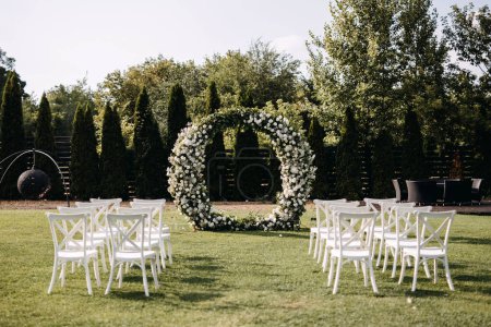 Téléchargez les photos : Allée de cérémonie de mariage avec une arche ronde décorée de fleurs et de chaises en bois blanc placées sur de l'herbe verte. Lieu de mariage jardin. - en image libre de droit