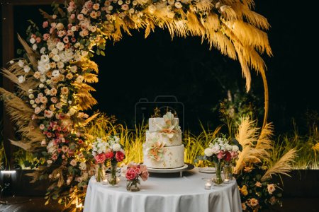 Photo for Three layered wedding cake at night, decorated with flowers made of frosting, on a round arch background. - Royalty Free Image