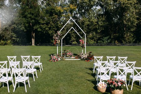 Photo for Wedding ceremony aisle with an arch decorated with flowers and white wooden chairs placed on green grass. Garden wedding venue. - Royalty Free Image