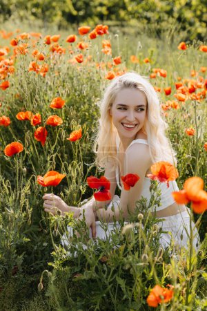 Téléchargez les photos : Joyeux jeune femme assise dans un champ avec des coquelicots rouges sauvages, souriant. - en image libre de droit