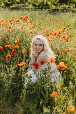Photo for Young woman sitting in a field with wild red poppies, smiling. - Royalty Free Image