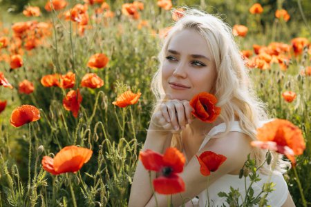 Photo for Portrait of a blonde young woman in a field with wild red poppies, smiling, holding a red poppy. - Royalty Free Image