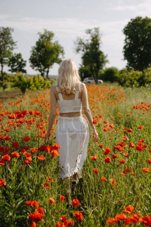 Foto de Mujer rubia caminando en un campo con amapolas rojas salvajes, usando un vestido blanco. - Imagen libre de derechos