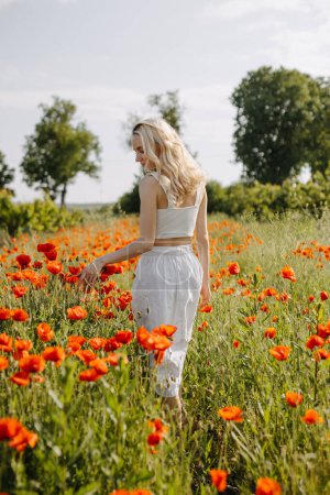 Téléchargez les photos : Jeune femme blonde debout dans un champ avec des coquelicots rouges sauvages, portant une robe blanche. - en image libre de droit