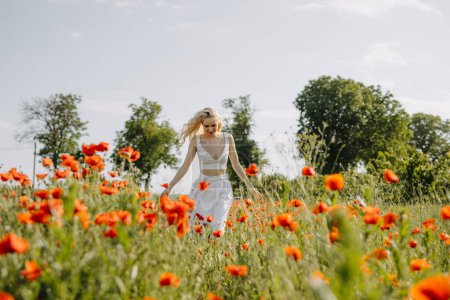 Téléchargez les photos : Femme blonde courant dans un champ avec des coquelicots rouges sauvages, portant une robe blanche. - en image libre de droit