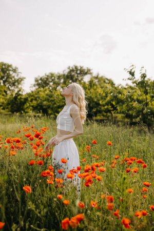 Téléchargez les photos : Femme blonde debout dans un champ avec des coquelicots rouges sauvages, portant une robe blanche, profitant de l'air frais. - en image libre de droit