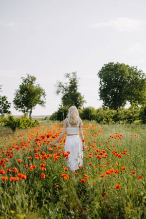 Téléchargez les photos : Femme blonde marchant dans un champ avec des coquelicots rouges sauvages, portant une robe blanche. - en image libre de droit