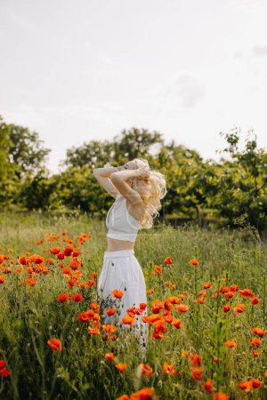 Téléchargez les photos : Femme blonde debout dans un champ avec des coquelicots rouges sauvages, portant une robe blanche. - en image libre de droit