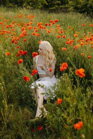 Téléchargez les photos : Femme blonde assise dans un champ avec des coquelicots rouges sauvages, portant une robe blanche, profitant de la nature. - en image libre de droit