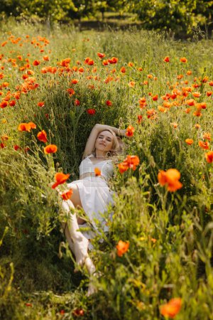 Téléchargez les photos : Femme couchée dans un champ avec des coquelicots rouges sauvages, portant une robe blanche, profitant de la nature. - en image libre de droit