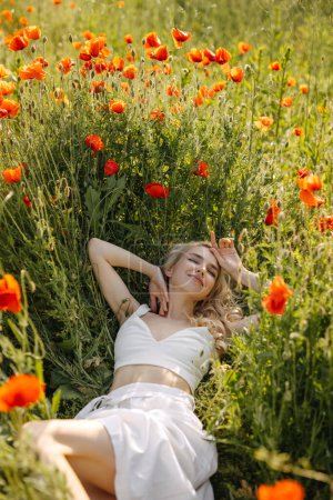 Téléchargez les photos : Heureuse femme blonde allongée dans un champ avec des coquelicots rouges sauvages, portant une robe blanche, profitant de la nature. - en image libre de droit