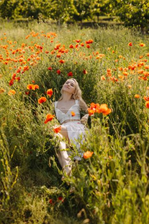 Téléchargez les photos : Femme couchée dans un champ avec des coquelicots rouges sauvages, portant une robe blanche, profitant de la nature. - en image libre de droit