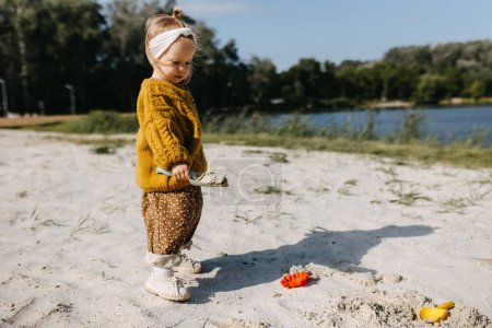 Téléchargez les photos : Petite fille jouant dans le sable, avec des moules de sable et une pelle, sur une plage au bord du lac. - en image libre de droit