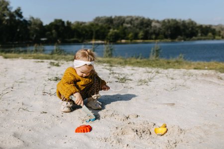 Téléchargez les photos : Petite fille jouant dans le sable, avec des moules de sable et une pelle, sur une plage au bord du lac. - en image libre de droit