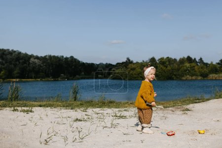 Foto de Niña jugando en la arena, con moldes de arena y una pala, en una playa junto al lago. - Imagen libre de derechos