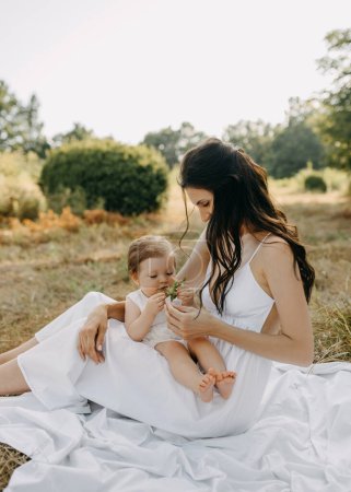 Photo for Mother holding little daughter on laps, playing with her, sitting on grass in a park on summer day. - Royalty Free Image