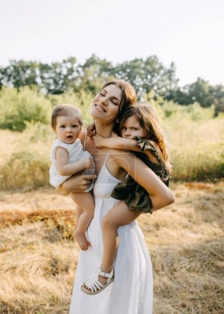 Photo for Mother holding her two daughters in arms, outdoors in a park on a summer day. - Royalty Free Image