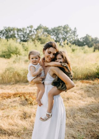 Photo for Mother playing with her two daughters outdoors in a park on a summer day, holding them in arms. - Royalty Free Image