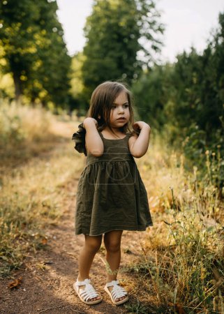 Téléchargez les photos : Petite fille sérieuse debout seule sur un chemin dans un parc, vêtue d'une robe de lin, le jour d'été. - en image libre de droit
