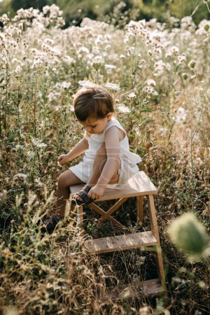 Photo for Little girl sitting on a wooden chair in a field, putting on her shoes. - Royalty Free Image