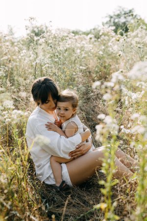 Téléchargez les photos : Mère embrassant petite fille, souriante, assise dans un champ avec de l'herbe haute. - en image libre de droit