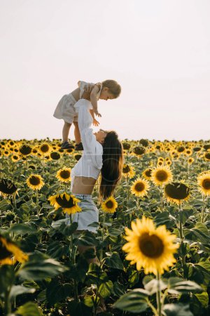 Téléchargez les photos : Mère jouant avec sa fille, la tenant vers le ciel, dans un champ sans fin de tournesols. - en image libre de droit