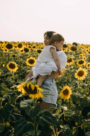 Téléchargez les photos : Mère debout avec sa petite fille à l'extérieur dans un champ de tournesol, la tenant dans ses bras. - en image libre de droit