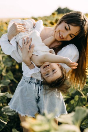Photo for Mother playing with her little daughter outdoors in a sunflower field, holding her in arms, laughing. - Royalty Free Image