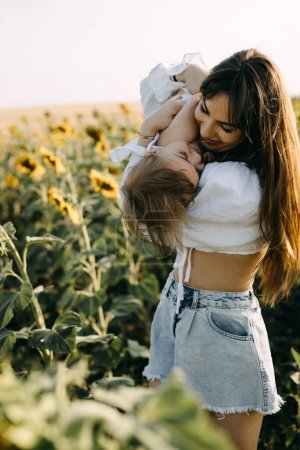Photo for Mother playing with her little daughter outdoors in a sunflower field, holding her in arms. - Royalty Free Image