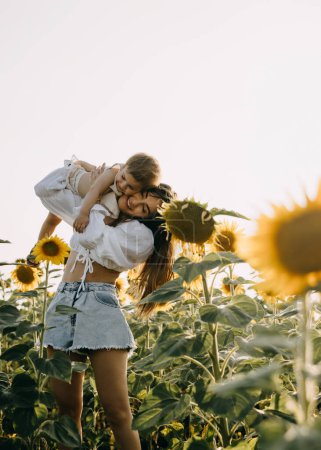 Photo for Mother playing with her little daughter outdoors in a sunflower field, holding her in arms. - Royalty Free Image