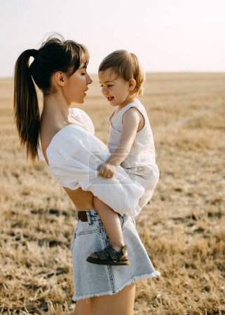 Téléchargez les photos : Mère tenant sa petite fille, debout dans un champ ouvert un jour d'été, vêtue de vêtements blancs. - en image libre de droit
