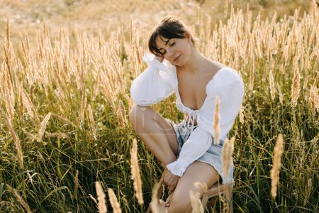 Téléchargez les photos : Jeune femme assise sur une chaise dans un champ avec de l'herbe sèche haute, les yeux fermés, rêvant, souriant. - en image libre de droit