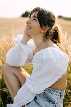 Foto de Feliz joven sentada en un campo con hierba seca alta, soñando, sonriendo. - Imagen libre de derechos