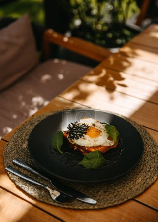 Téléchargez les photos : Œuf frit sur pain grillé, sur une assiette noire sur une table en bois sur une terrasse. - en image libre de droit