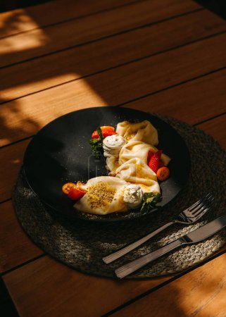 Téléchargez les photos : Crêpes à la crème et fraises sur une assiette noire au soleil. - en image libre de droit