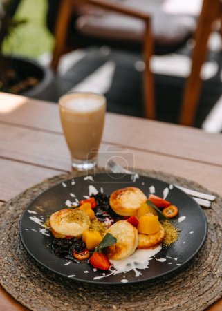 Téléchargez les photos : Crêpes au fromage cottage avec fruits sur assiette et latte sur table en bois dans un café terrasse en plein air. - en image libre de droit