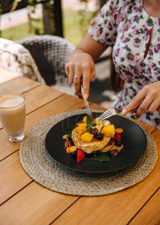 Téléchargez les photos : Gros plan d'une femme mangeant des crêpes garnies de fruits et de noix, servie avec un latte sur une table en bois. - en image libre de droit