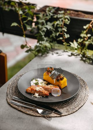 Foto de Polenta y carne con una salsa cremosa en un plato negro, entorno al aire libre en una terraza cafetería. - Imagen libre de derechos