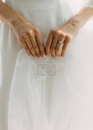 Téléchargez les photos : Les mains d'une femme avec une bague de fiançailles tenant doucement un voile sur le fond de la robe de mariée. - en image libre de droit