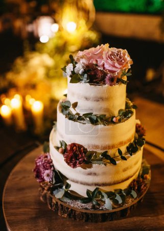 Téléchargez les photos : Un gâteau de mariage à trois niveaux orné de fleurs fraîches et de verdure, sur fond de lumière chaude aux chandelles. - en image libre de droit