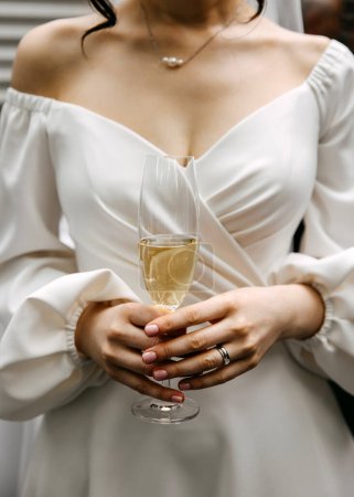 Foto de Una novia en un elegante vestido blanco fuera del hombro sosteniendo una flauta de champán, con un anillo de diamantes. - Imagen libre de derechos