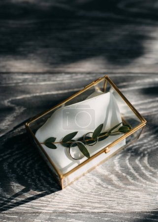 Foto de Caja de vidrio con anillos de boda en un paño de seda con sombras dramáticas de fundición de luz en una superficie de madera. - Imagen libre de derechos